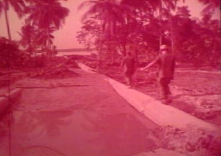 Screenshot from original film of two men walking on a pipe at the Dong Tam Base Camp Construction. Image is red tinted.