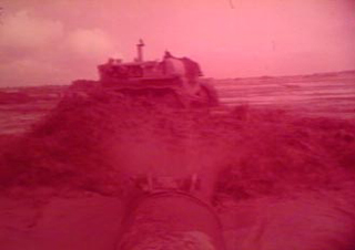 Screenshot from the original film of a pipe spraying water and a bulldozer moving dirt during the construction of the camp. Image is red tinted.